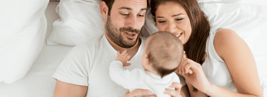 Couple qui cajole son bébé allongés dans le lit - L’arrivée d’un enfant et les difficultés pour le couple! - Blogue | Clinique de Psychologie Québec